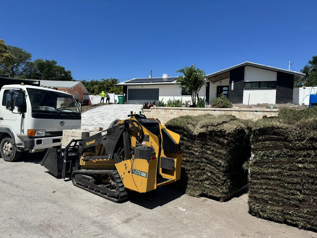 Landscaping crew installing fresh sod and a paver driveway at a home, with a compact track loader and pallets of turf
